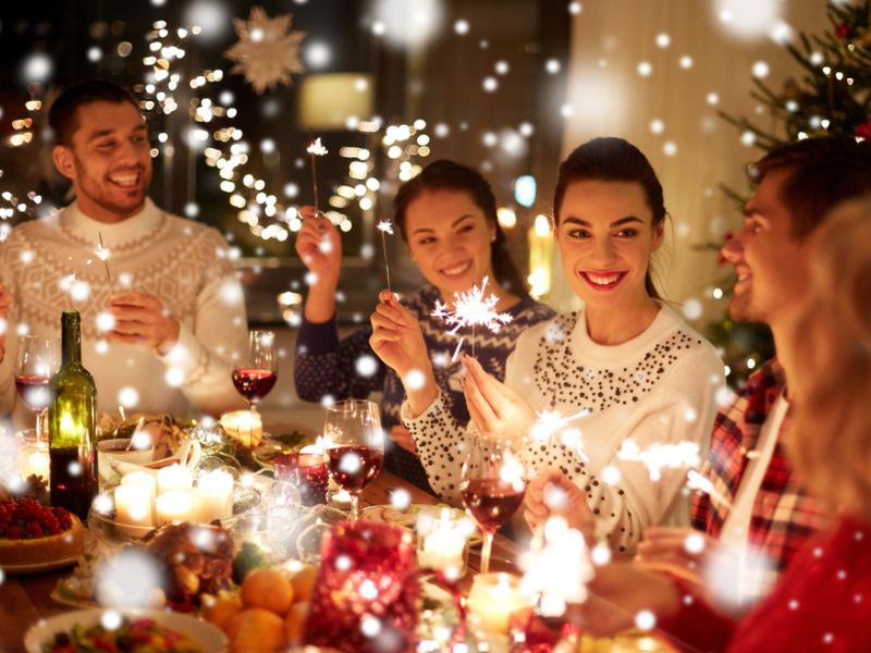 A group of friends celebrating indoors around a festive dinner table, holding sparklers and smiling, surrounded by warm holiday lights and decorations